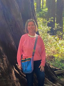 Kristen Brown, a white woman wearing a pink shirt, is standing in front of a large tree, with sunny green leaves in the background. She has short light-colored hair and she is smiling.